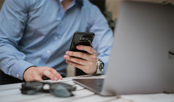 Man in a blue shirt using a smartphone while working on a laptop at a desk.